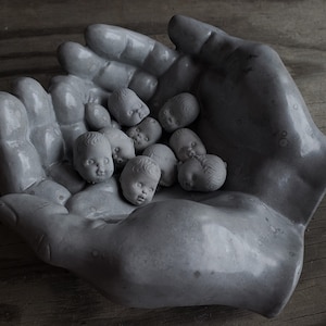 May include: A gray concrete hand sculpture holding a collection of small gray baby doll heads.