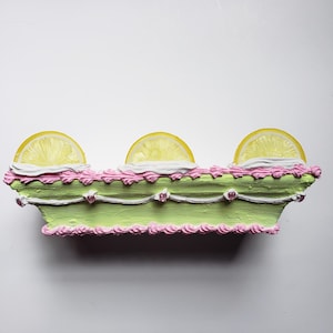Puede incluir: Una pieza decorativa rectangular con base verde claro, glaseado rosa y tres rodajas de limón. La pieza tiene adornos decorativos blancos y rosas y pequeños adornos en forma de corazón. El diseño se asemeja a un pastel.
