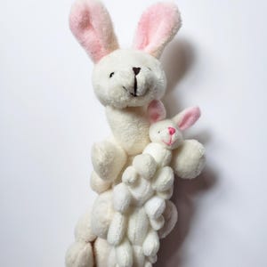 May include: A stack of white plush bunny toys with pink ears. The top bunny is the largest and has a smaller bunny nestled in its arms.