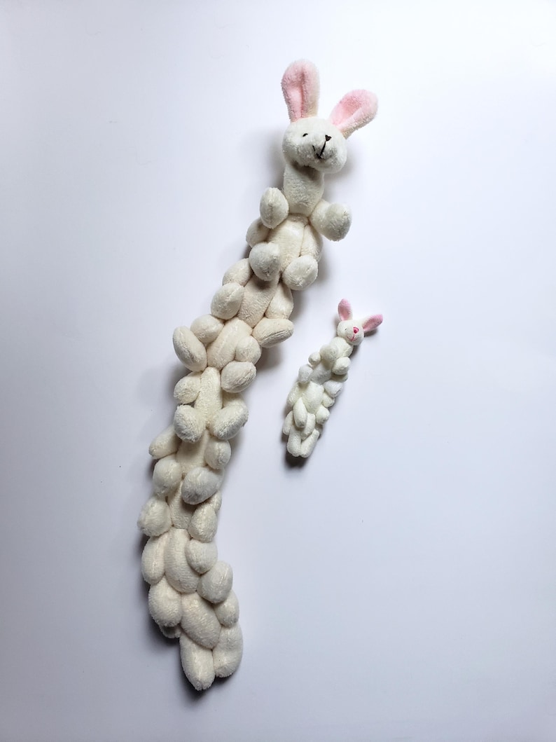 May include: A white plush bunny toy with pink ears is stacked on top of a long chain of smaller white plush bunnies. The chain of bunnies is arranged in a vertical line.