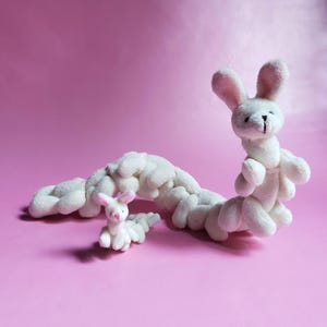 May include: A white plush bunny toy shaped like a caterpillar. The bunny has pink ears and a pink nose. The bunny is lying on a pink surface.