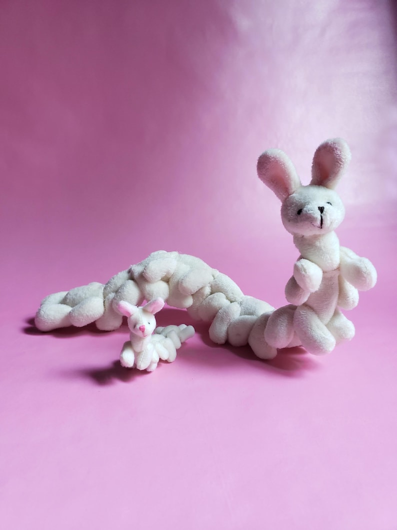 May include: A white plush bunny toy shaped like a caterpillar. The bunny has pink ears and a pink nose. The bunny is lying on a pink surface.