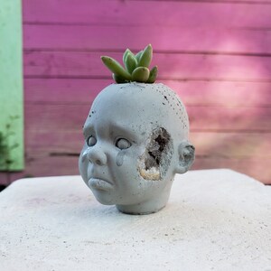 May include: A grey concrete planter in the shape of a crying baby's head, with a hole in the side revealing a crystal. A small green succulent grows from the top. The planter is on a white surface.