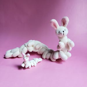 May include: Two white plush bunny toys in the shape of a caterpillar. The larger bunny has a pink nose and ears. The smaller bunny is curled up in a ball.