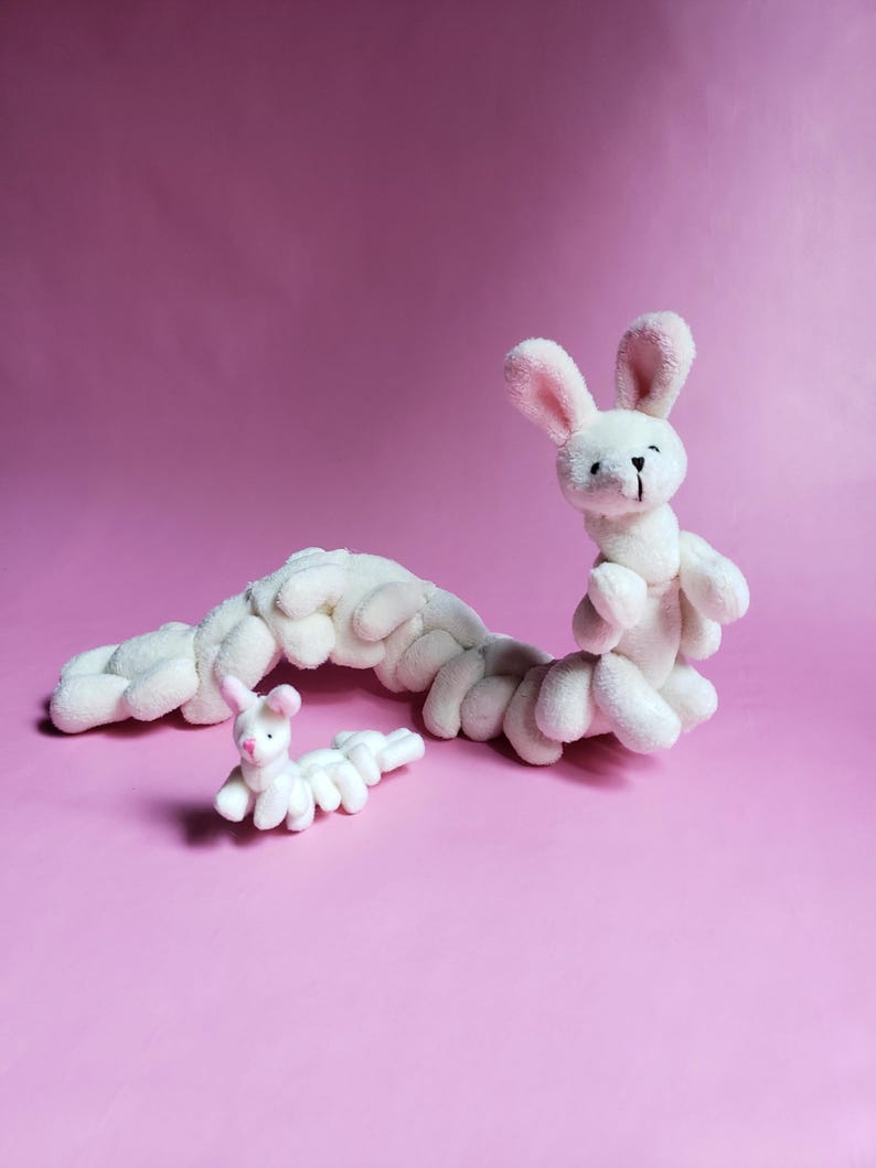 May include: Two white plush bunny toys in the shape of a caterpillar. The larger bunny has a pink nose and ears. The smaller bunny is curled up in a ball.