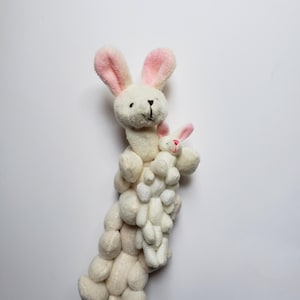 May include: A white plush bunny with pink ears holding a smaller white plush bunny with pink ears. The bunnies are stacked on top of each other, creating a tower of plush bunnies.