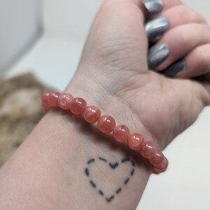 May include: A pink gemstone bracelet with a heart tattoo on a person's wrist.