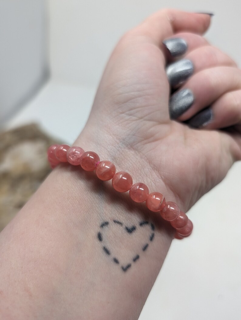 May include: A pink gemstone bracelet with a heart tattoo on a person's wrist.