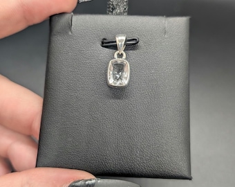 May include: A silver pendant with a rectangular, clear gemstone is displayed on a black presentation card. The pendant is suspended from a black cord. The background is a solid black.