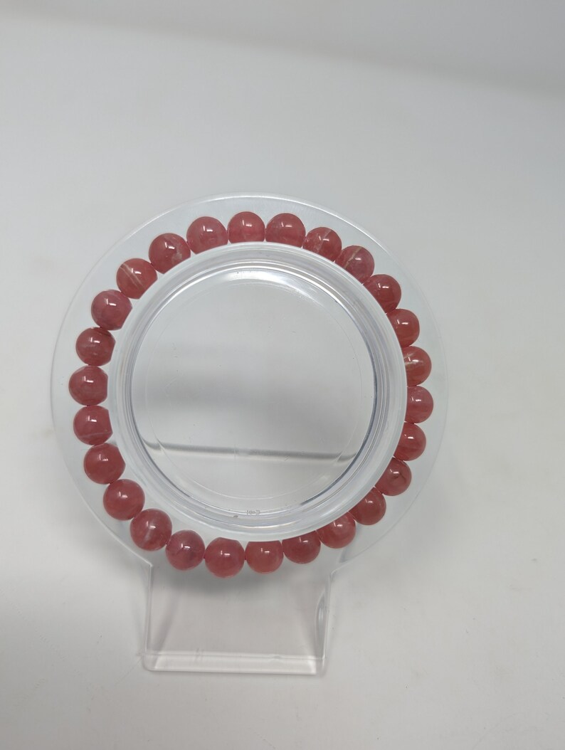 May include: A pink and white gemstone bracelet with round beads. The bracelet is on a clear plastic stand.