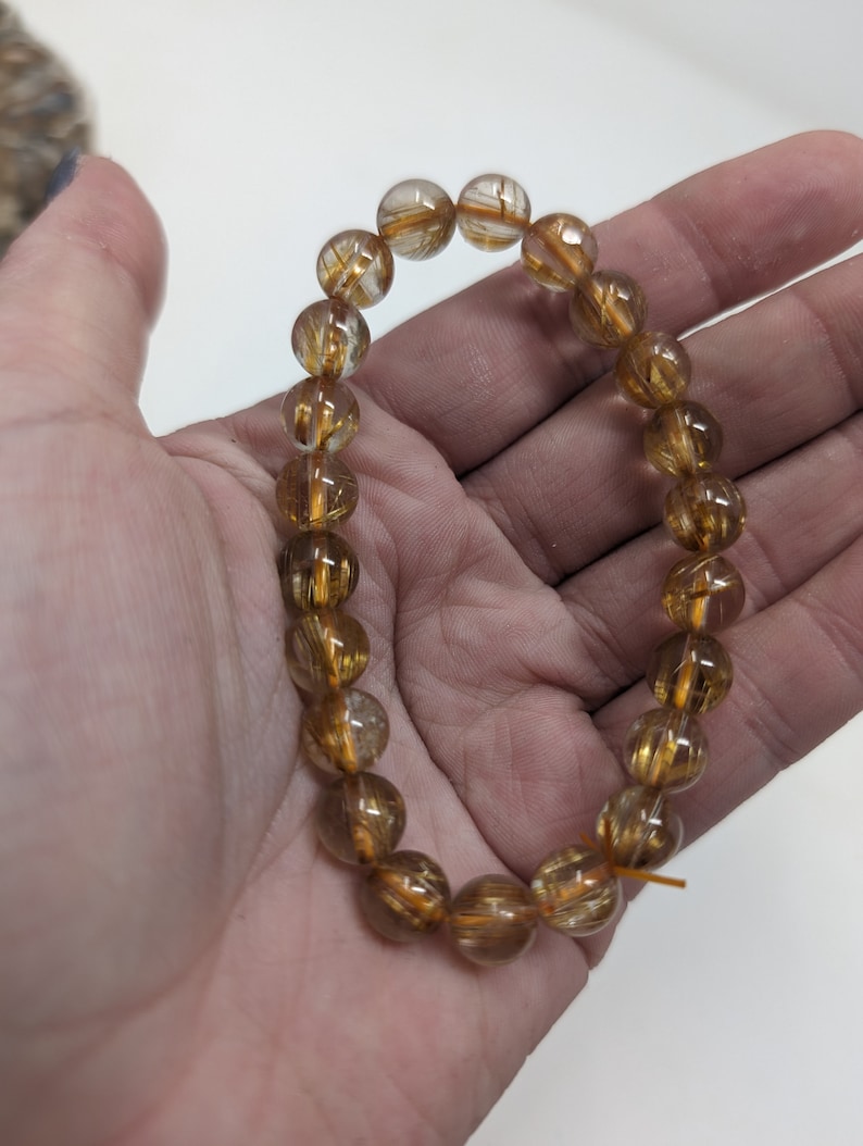 May include: A gold and clear quartz bracelet with gold inclusions. The bracelet is made of round beads and is strung on a clear elastic cord.