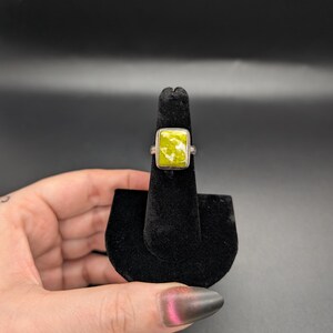 May include: A silver ring with a square, light green stone. The ring is displayed on a black velvet ring holder against a dark background. The stone has a textured appearance.