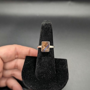 May include: A silver ring with a rectangular stone featuring brown and blue patterns. The ring is displayed on a black velvet ring holder against a dark background. The stone's unique color and pattern are the focal point.