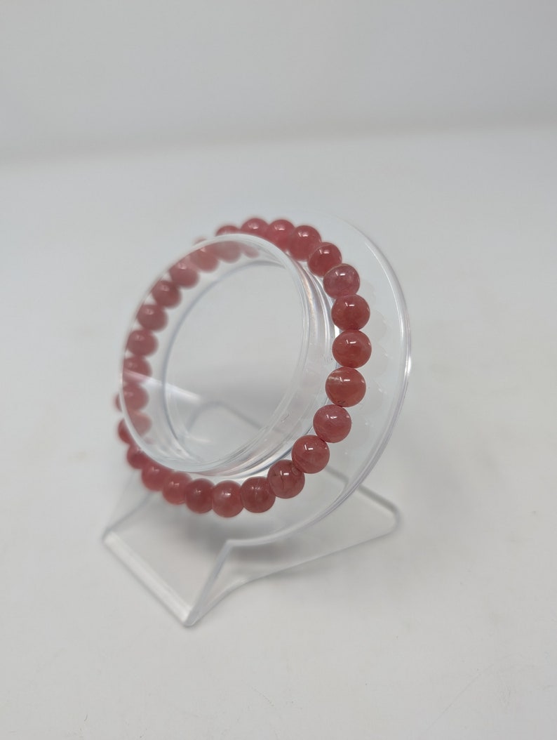 May include: A pink gemstone bracelet with round beads on a clear display stand.