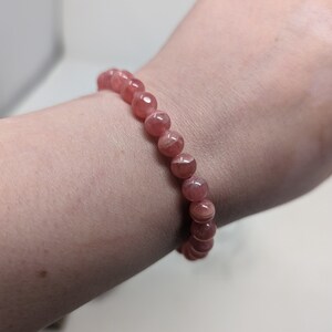 May include: A pink gemstone bracelet with round beads. The bracelet is on a person's wrist.