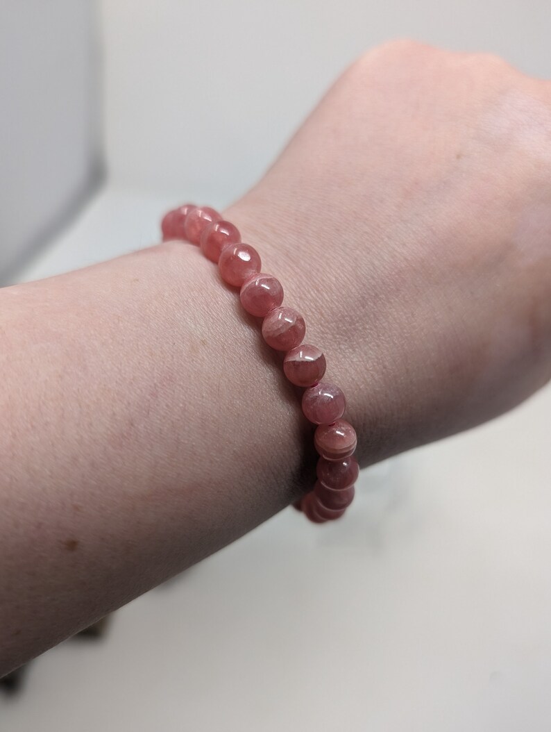 May include: A pink gemstone bracelet with round beads. The bracelet is on a person's wrist.