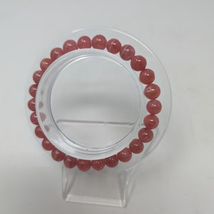 May include: A pink gemstone bracelet with round beads. The bracelet is displayed on a clear plastic stand.