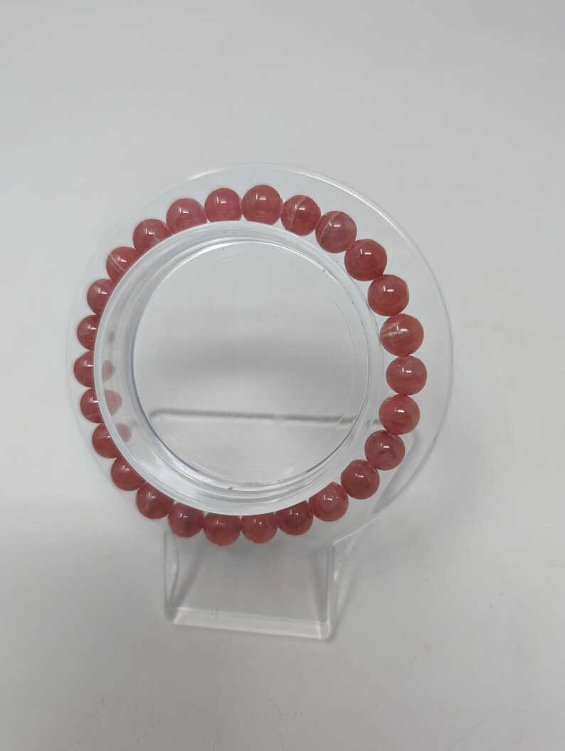 May include: A pink gemstone bracelet with round beads. The bracelet is displayed on a clear plastic stand.