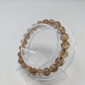 May include: A clear quartz bracelet with gold inclusions. The bracelet is made of round beads and is displayed on a clear plastic stand.