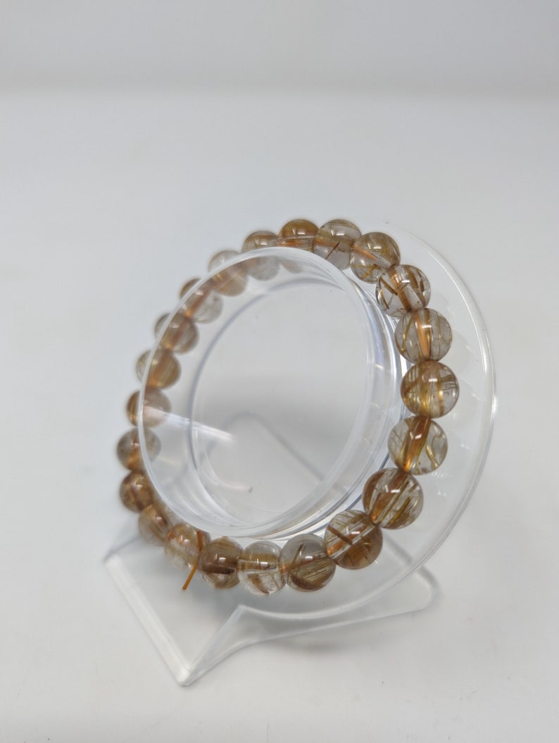 May include: A clear quartz bracelet with gold inclusions. The bracelet is made of round beads and is displayed on a clear plastic stand.