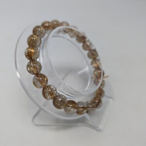May include: A gold and clear quartz crystal bracelet with a wire-like inclusion. The bracelet is on a clear plastic stand.