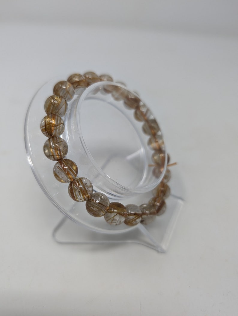 May include: A gold and clear quartz crystal bracelet with a wire-like inclusion. The bracelet is on a clear plastic stand.