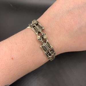 May include: A gold-toned bracelet featuring a repeating pattern of two skeletons holding hands. The skeletons are facing each other and have detailed features, including skulls and ribs.