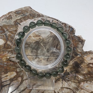 May include: A green gemstone bracelet with round beads on a clear display stand. The bracelet is on a brown and white background.