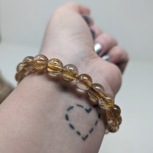 May include: A gold and clear crystal beaded bracelet with a heart tattoo on the wrist.