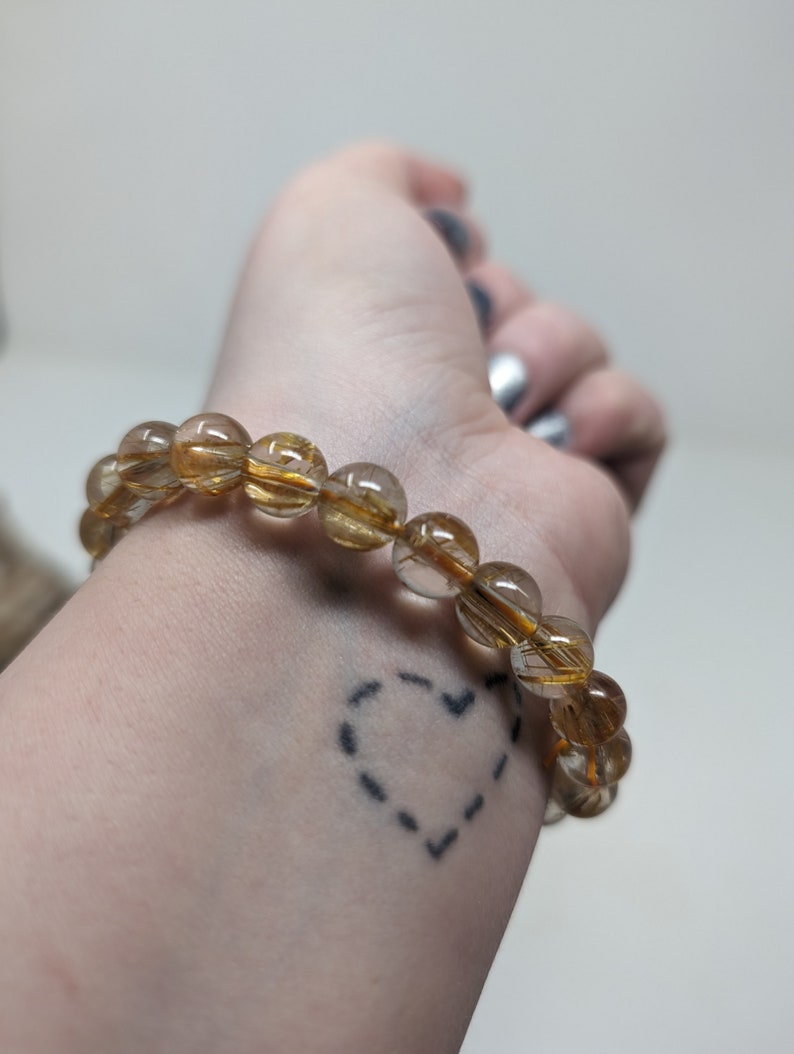 May include: A gold and clear crystal beaded bracelet with a heart tattoo on the wrist.