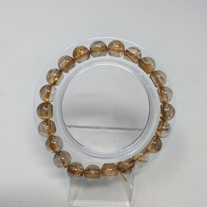 May include: A gold and clear crystal bracelet with gold flecks throughout. The bracelet is made of round beads and is displayed on a clear plastic stand.