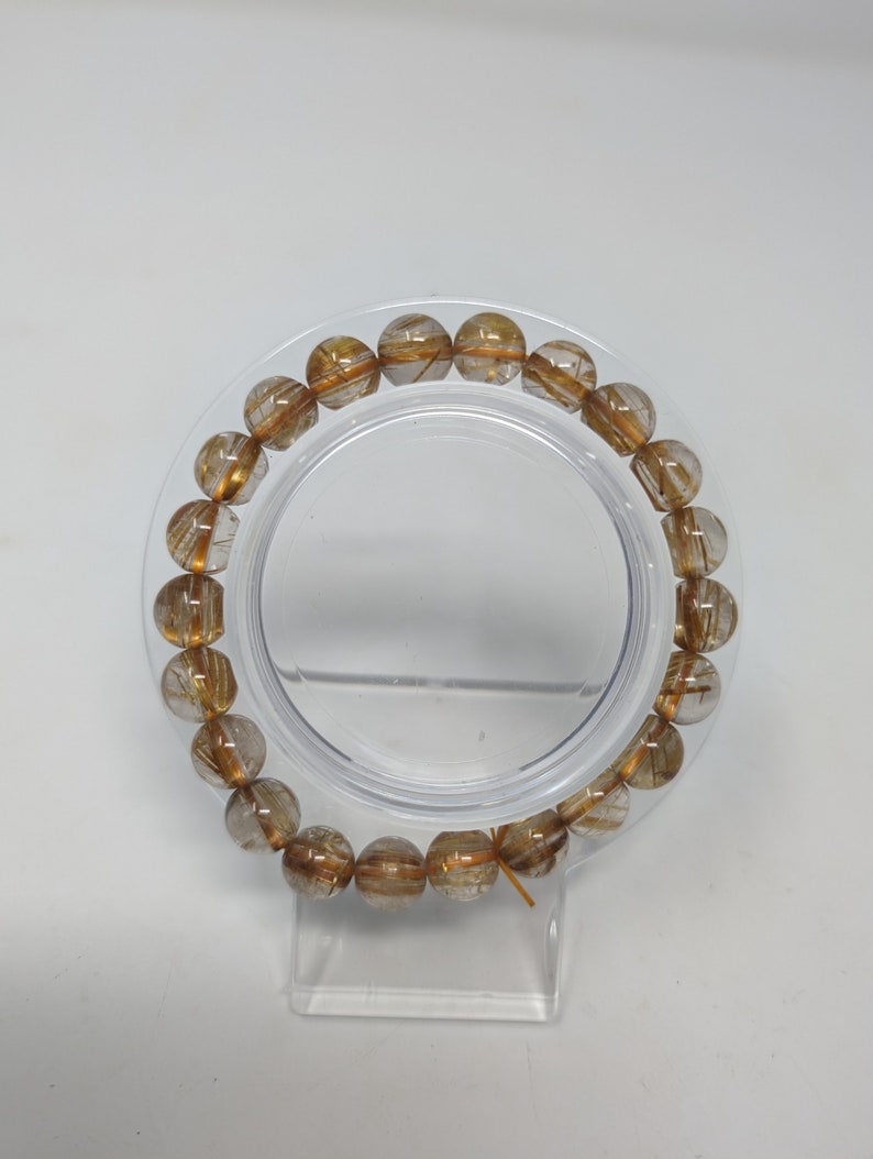 May include: A gold and clear crystal bracelet with gold flecks throughout. The bracelet is made of round beads and is displayed on a clear plastic stand.