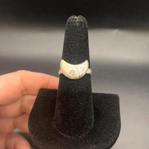 May include: A silver ring with a white crescent moon design. The ring is on a black ring display stand.