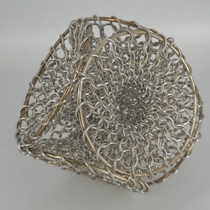 May include: A silver and gold metal sculpture made of chainmail. The sculpture is shaped like a sphere with a hole in the center.