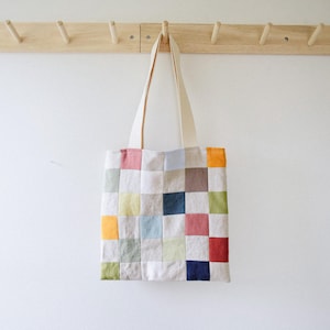 Peut inclure: Un sac fourre-tout patchwork avec une poignée crème. Le corps du sac est une grille de carrés colorés dans les tons de rose, bleu, jaune, vert et orange. Le sac est suspendu à un porte-manteau en bois avec des chevilles.
