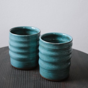 May include: Two cylindrical ceramic tumblers featuring a vibrant teal glaze with a glossy finish. The exterior of each vessel displays a series of horizontal, raised ridges that create a textured, undulating surface around the body of the cups.