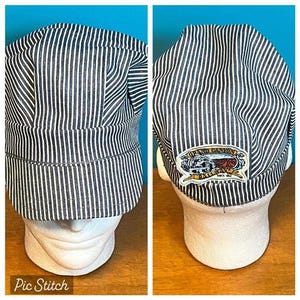 May include: A blue and white striped conductor hat with a Grand Canyon patch. The hat is displayed on a white mannequin head. The hat has a short brim and a rounded top. The patch features a train and the words "Grand Canyon".