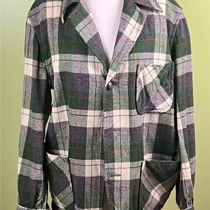 K&ouml;nnte beinhalten: Eine gr&uuml;n, grau und cremefarbene karierte Jacke mit Kragen, drei Vordertaschen und Knopfverschl&uuml;ssen. Die Jacke wird auf einer Schaufensterpuppe pr&auml;sentiert und hat lange &Auml;rmel.