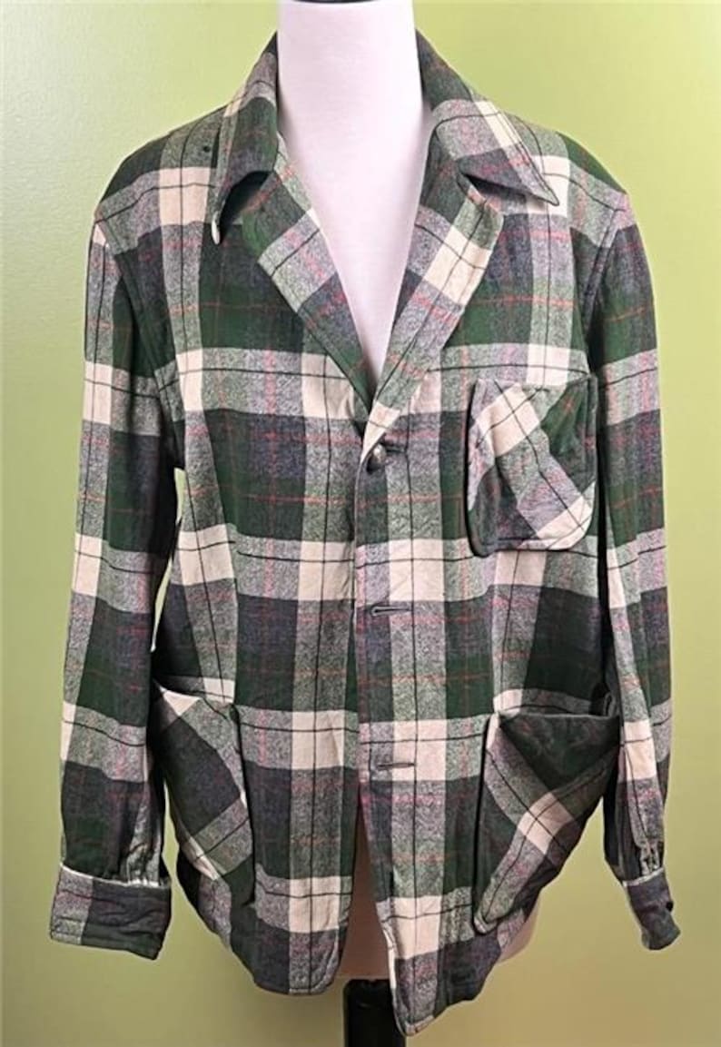 K&ouml;nnte beinhalten: Eine gr&uuml;n, grau und cremefarbene karierte Jacke mit Kragen, drei Vordertaschen und Knopfverschl&uuml;ssen. Die Jacke wird auf einer Schaufensterpuppe pr&auml;sentiert und hat lange &Auml;rmel.