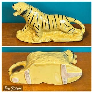 May include: A ceramic tiger figurine in a reclining pose, painted yellow with black stripes. The tiger is resting on a textured base, with its mouth open. The figurine is a decorative item.