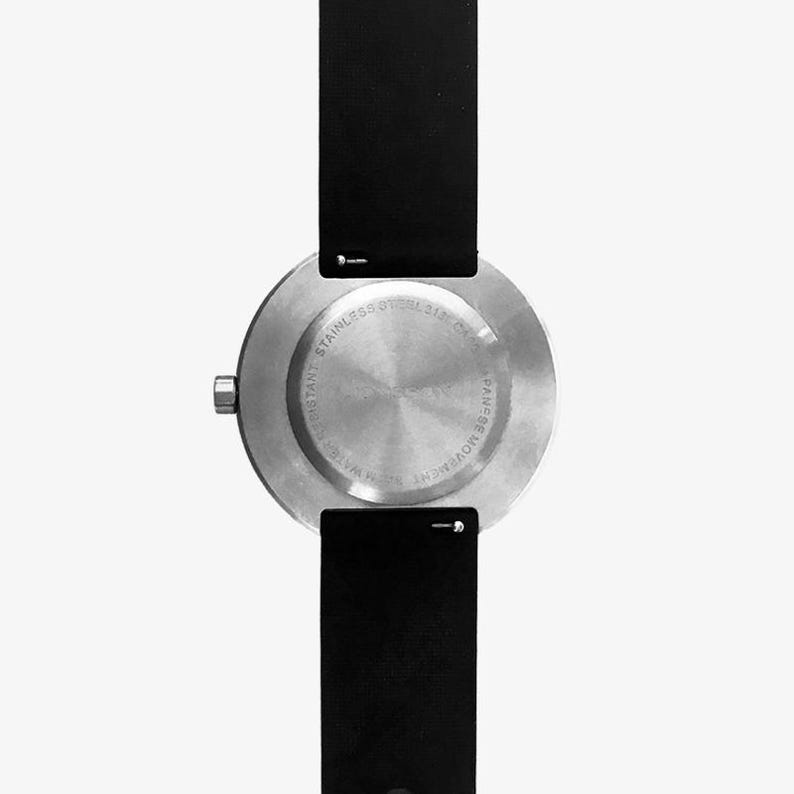 May include: The back of a silver watch with a black leather band. The watch face has the text "STAINLESS STEEL 316L CASE" and "JAPAN MOVEMENT" printed on it.
