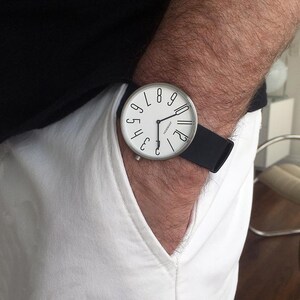 May include: A white wristwatch with a black leather band. The watch face has black numbers 1 through 12 arranged in a circle around the center. The watch face also has the word "Norsewood" printed in black.