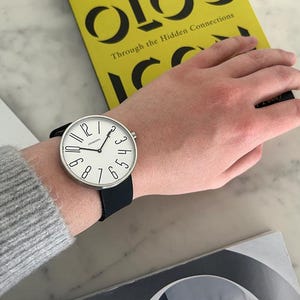 May include: A silver wristwatch with a white face and black leather band. The watch face has black numbers and a black second hand. The watch is on a person's wrist.