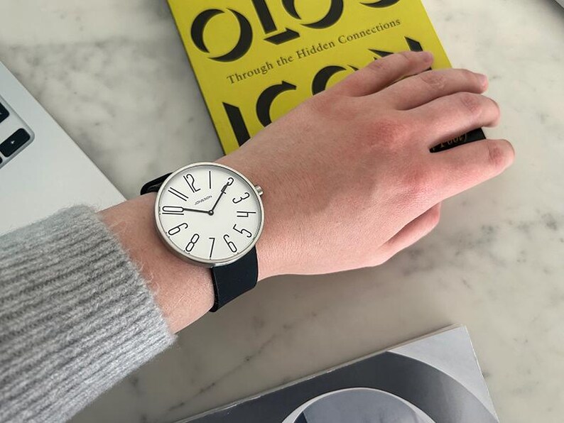 May include: A silver wristwatch with a white face and black leather band. The watch face has black numbers and a black second hand. The watch is on a person's wrist.