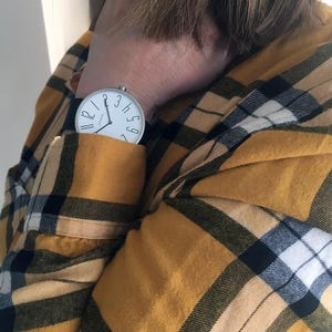 May include: A white analog wristwatch with black hands and black numbers on a black leather band. The watch is worn on a person's wrist, with the watch face facing the camera. The person is wearing a yellow and black plaid shirt.