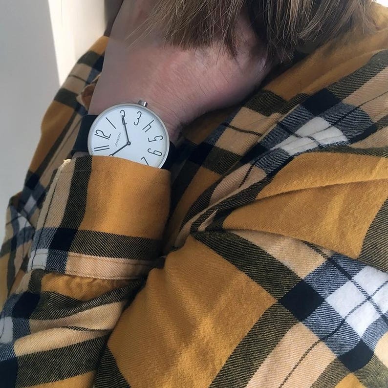 May include: A white analog wristwatch with black hands and black numbers on a black leather band. The watch is worn on a person's wrist, with the watch face facing the camera. The person is wearing a yellow and black plaid shirt.