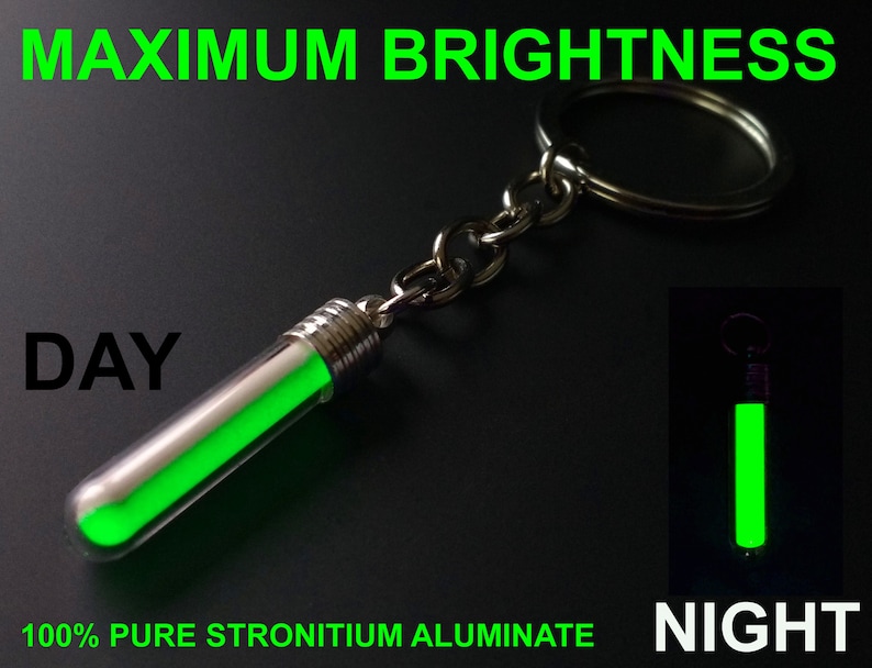 Puede incluir: Un colgante de llavero verde brillante en un tubo transparente, con una tapa y cadena de metal plateado. El texto "MAXIMUM BRIGHTNESS" y "100% PURE STRONTIUM ALUMINATE" se muestran en verde. La imagen tambi&eacute;n muestra el colgante brillando por la noche.