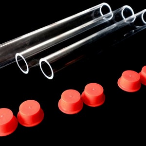 Puede incluir: Tres tubos de vidrio transparente con extremos redondeados se colocan sobre seis tapones de goma rojos. Los tubos son cilíndricos y transparentes. Los tapones son de color rojo brillante y tienen forma cónica.