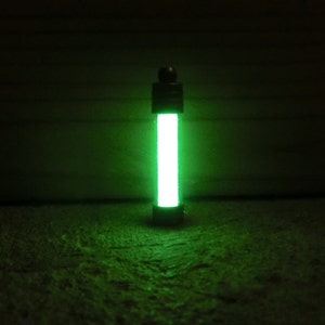 Puede incluir: Un objeto cil&iacute;ndrico verde brillante con una tapa y base oscuras. El objeto emite una luz verde brillante y vibrante, creando un contraste llamativo con el fondo oscuro. El efecto general es una est&eacute;tica futurista o cient&iacute;fica.