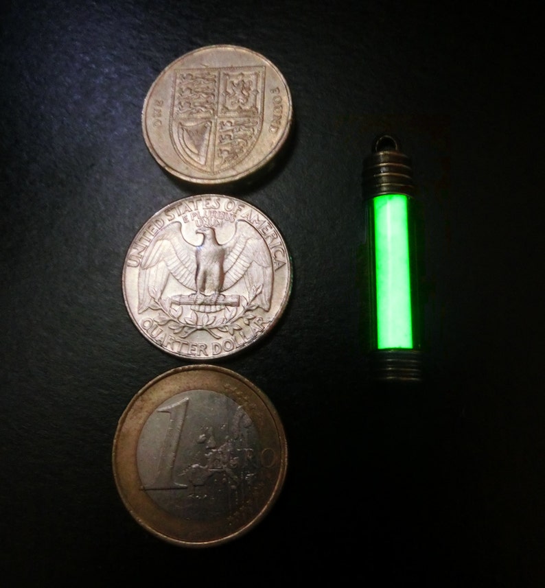 Puede incluir: Un colgante verde brillante junto a una moneda de euro, una moneda de veinticinco centavos de d&oacute;lar estadounidense y una moneda de una libra esterlina brit&aacute;nica. El colgante es un tubo peque&ntilde;o y transparente con un interior verde brillante. Las monedas son plateadas y doradas.